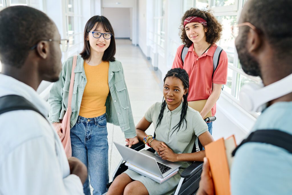 African American teenage girl with disability sitting on wheelchair with laptop on her knees and talking to her friends