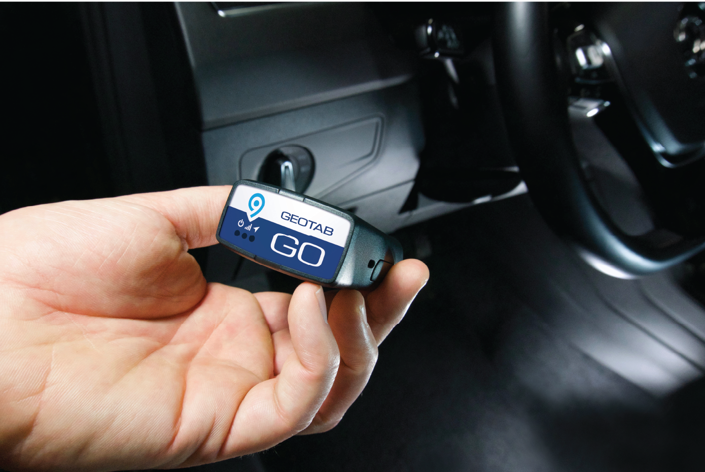 a hand holding a Geotab GO device, which is used for vehicle tracking and telematics. The device, with its label visible, is being prepared for installation inside a vehicle, most likely by plugging it into the vehicle’s diagnostic port. This type of device is used in fleet management to monitor data such as location, driving behavior, and vehicle diagnostics, helping businesses track and manage their vehicles in real-time. The vehicle's dashboard and steering wheel are partially visible in the background.