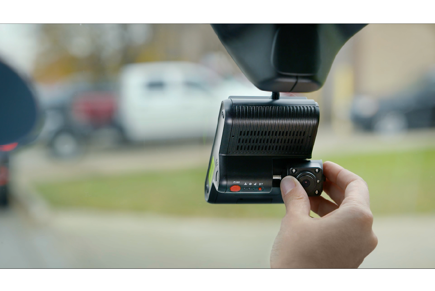 a person adjusting a dashboard-mounted camera inside a vehicle. The camera appears to be a dashcam or telematics device used to monitor driving conditions or driver behavior. The hand is shown rotating a knob on the device, which is mounted near the rearview mirror, with a blurred background of vehicles and greenery outside the window.