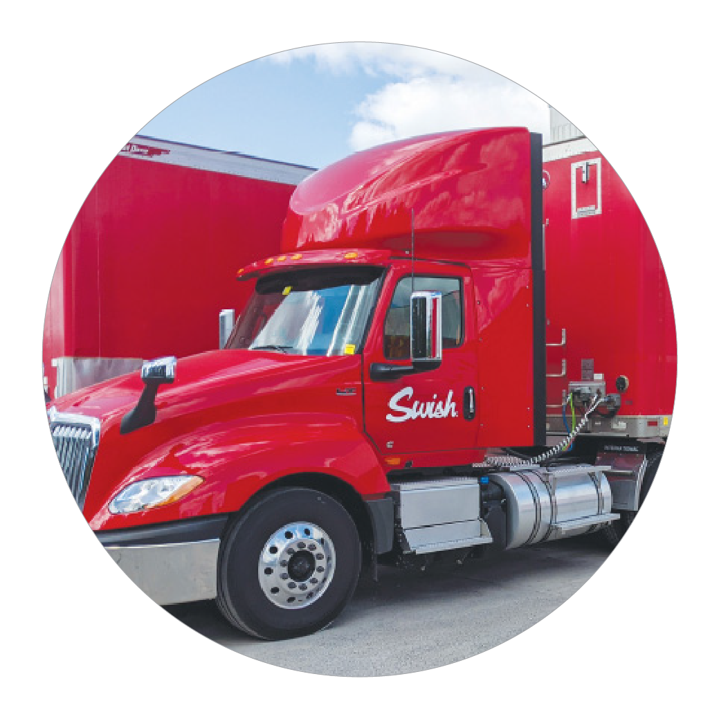 a large red Swish-branded truck parked at a loading dock, with a trailer displaying the text "Canada's source for quality cleaning supplies and equipment" along with the company's website, swish.ca. The truck is attached to a matching trailer, emphasizing the company's fleet for logistics and delivery. The bright red color and the large logo make the vehicle and brand highly visible, highlighting Swish's role in distributing cleaning supplies. The scene suggests an active distribution center focused on efficient transportation.