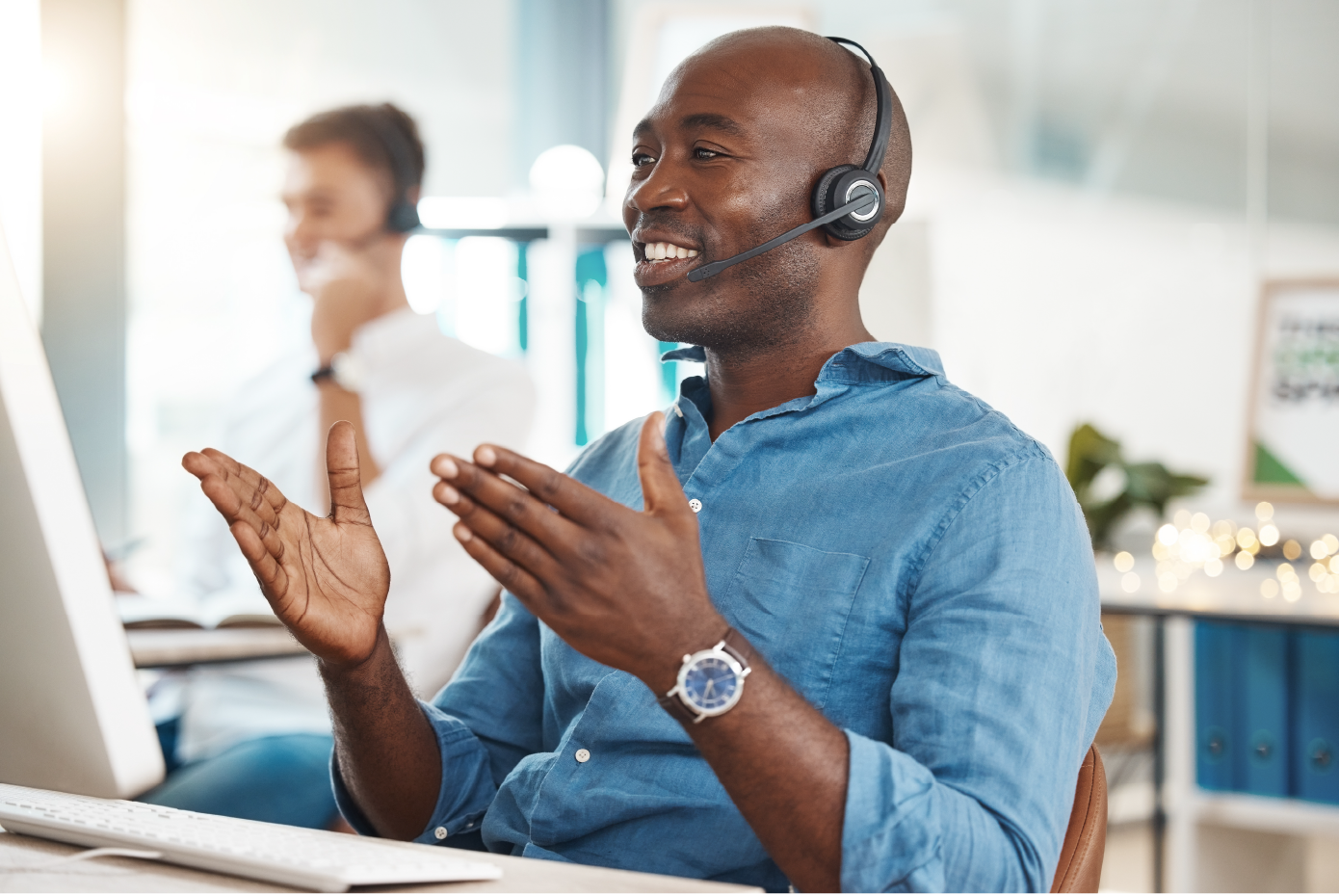 person wearing a headset and gesturing with their hands while smiling, likely in a customer service or call center setting. The individual appears to be engaged in a conversation or offering assistance, with a background showing another person also wearing a headset, suggesting a busy office environment.