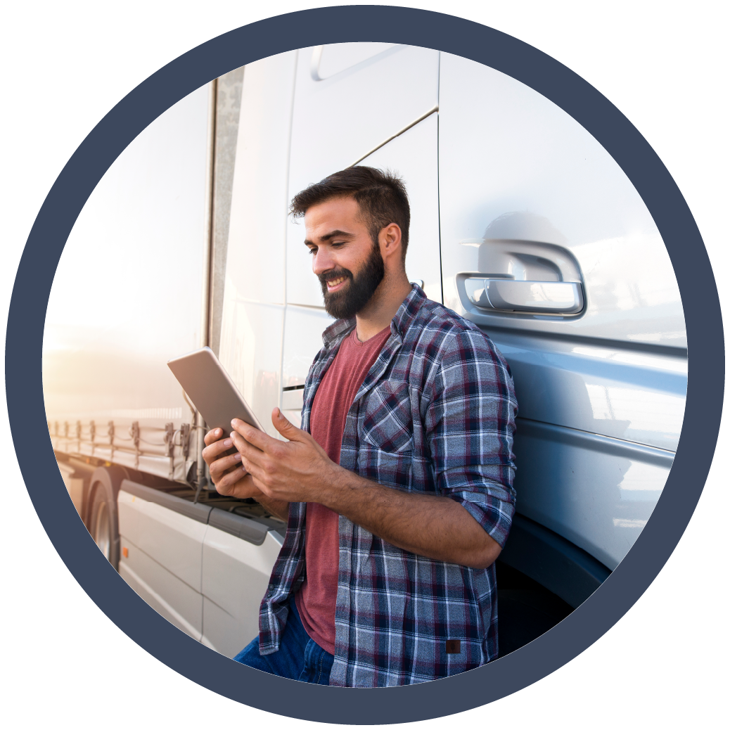 a man leaning against a large truck while holding and using a tablet. He is wearing a casual plaid shirt and smiling, suggesting he is either reviewing information or communicating via the tablet. The truck and the outdoor setting indicate that this may be related to logistics, trucking, or fleet management, highlighting the use of technology in modern transportation operations.