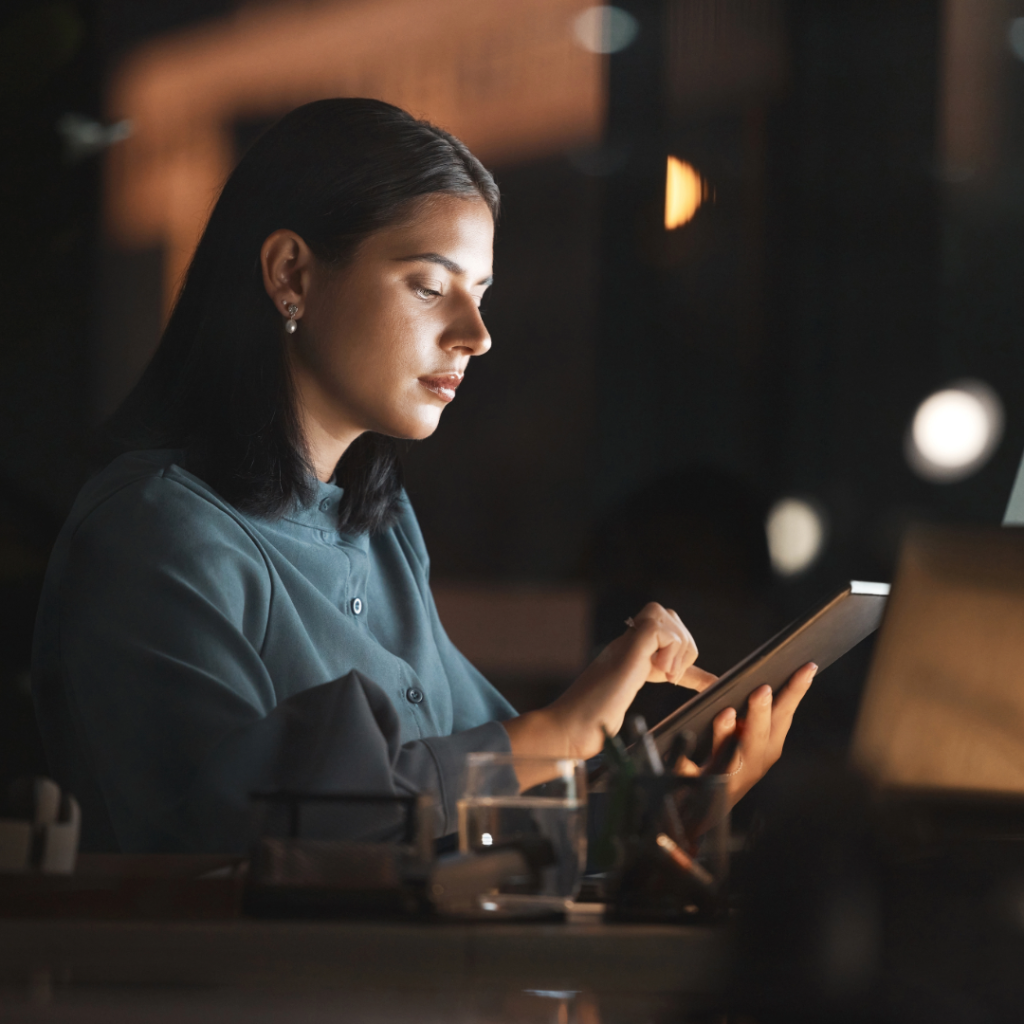 A woman sits at her desk in a dimly lit office, deeply focused on a tablet. The surrounding environment, with subtle lighting and a glass of water on the desk, conveys a sense of concentration and dedication, suggesting late-night work or analysis.