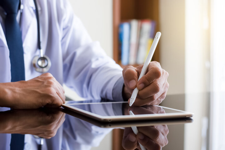 A close-up image of a doctor wearing a white coat with a stethoscope around their neck, using a stylus on a tablet. The reflection of the device on a polished surface emphasizes modern digital record-keeping in a professional healthcare setting.